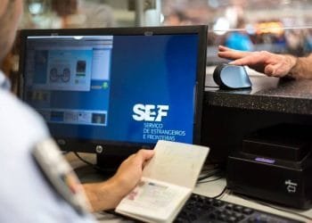 Per capire cosa è il lavoro di un agente di frontiera che tiene in mano un passaporto mentre digita su un computer. Il monitor visualizza SEF. È visibile la mano di un passeggero, appoggiata su uno scanner di impronte digitali. L'ambientazione sembra essere un posto di controllo dell'immigrazione.