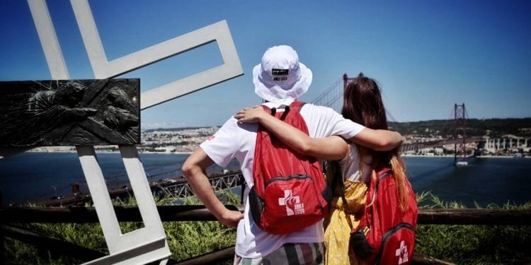 Due persone con zaini e cappelli da sole stanno di fronte a una vista panoramica di un ponte e dello skyline della città attraverso un corso d'acqua. Si abbracciano e osservano la vista. In primo piano c'è una grande croce bianca e su di essa è montata una scultura più piccola in rilievo scuro, che simboleggia la Giornata Mondiale Gioventù.