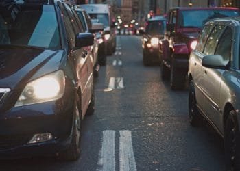 Una strada cittadina congestionata piena di auto bloccate nel traffico, ognuna con l'obbligo contrassegno assicurativo. I fari dei veicoli sono accesi e la strada è fiancheggiata da edifici a più piani. La scena cattura l'affollato ambiente urbano durante quella che sembra essere l'ora di punta.