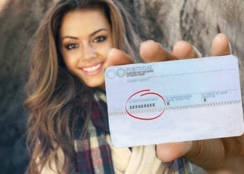 Una donna con i capelli lunghi e una sciarpa scozzese tiene in mano una tessera del cittadino portoghese verso la telecamera. La scheda mostra la sua foto e le informazioni personali, sebbene il numero di identificazione sia oscurato da una X per motivi di privacy. Se devi ottenere NIF Portogallo, avere questa carta è essenziale. Lo sfondo è sfocato.