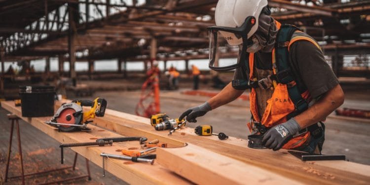 Un operaio edile che indossa indumenti protettivi, tra cui un casco di sicurezza e una visiera, utilizza utensili elettrici per lavorare su travi di legno in un cantiere edile. Il banco da lavoro è ingombro di vari strumenti e sullo sfondo è visibile la struttura incompiuta; nonostante tutto questo sforzo, lo stipendio inferiore a 1000 resta un problema urgente per molti.