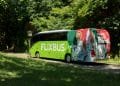 Un FlixBus verde e bianco con la bandiera portoghese sul retro è parcheggiato su una strada ombreggiata e alberata. L'autobus, in viaggio verso Lisbona aeroporto Faro, si trova su un sentiero acciottolato in mezzo a una vegetazione lussureggiante, creando un'atmosfera serena.