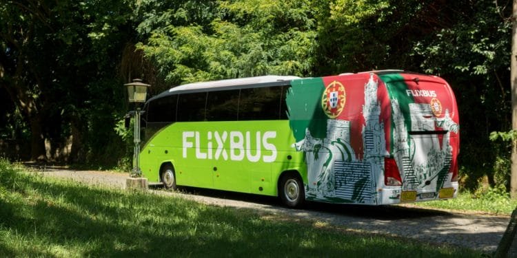 Un FlixBus verde e bianco con la bandiera portoghese sul retro è parcheggiato su una strada ombreggiata e alberata. L'autobus, in viaggio verso Lisbona aeroporto Faro, si trova su un sentiero acciottolato in mezzo a una vegetazione lussureggiante, creando un'atmosfera serena.