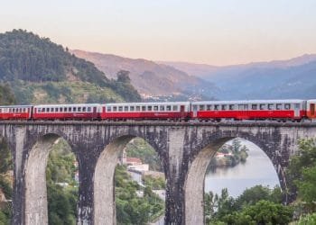 Un treno rosso e grigio viaggia su un lungo viadotto in pietra con archi multipli. Sotto, un fiume si snoda attraverso un paesaggio verde lussureggiante con montagne sullo sfondo sotto un cielo tenue dai colori pastello, incarnando il fascino del Portogallo, un percorso panoramico perfetto per chi ama viaggiare.