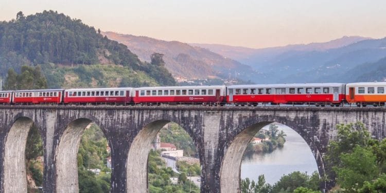 Un treno rosso e grigio viaggia su un lungo viadotto in pietra con archi multipli. Sotto, un fiume si snoda attraverso un paesaggio verde lussureggiante con montagne sullo sfondo sotto un cielo tenue dai colori pastello, incarnando il fascino del Portogallo, un percorso panoramico perfetto per chi ama viaggiare.