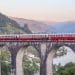 Un treno rosso e grigio viaggia su un lungo viadotto in pietra con archi multipli. Sotto, un fiume si snoda attraverso un paesaggio verde lussureggiante con montagne sullo sfondo sotto un cielo tenue dai colori pastello, incarnando il fascino del Portogallo, un percorso panoramico perfetto per chi ama viaggiare.