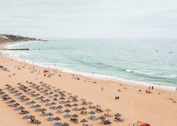 Un'ampia spiaggia sabbiosa del Caro Estate Algarve presenta file di sedie a sdraio e ombrelloni vicino alla riva. Le persone sono sparse sulla spiaggia, alcuni nuotano nel mare. Il cielo è coperto e sullo sfondo sono visibili alcuni edifici mentre le onde si infrangono dolcemente sulla riva.