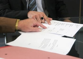 Due persone sono sedute a un tavolo di vetro e esaminano documenti. Una persona, che indossa un orologio d'oro e una manica marrone, indica il testo sui giornali, discutendo i dettagli di un conto corrente banca portoghese, mentre l'altra persona che indossa un anello nero e una giacca con cravatta a righe, segue la discussione.