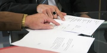 Due persone sono sedute a un tavolo di vetro e esaminano documenti. Una persona, che indossa un orologio d'oro e una manica marrone, indica il testo sui giornali, discutendo i dettagli di un conto corrente banca portoghese, mentre l'altra persona che indossa un anello nero e una giacca con cravatta a righe, segue la discussione.