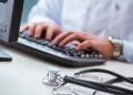 Una persona che indossa un camice bianco digita su una tastiera con uno stetoscopio posizionato sulla scrivania accanto a lei. La scena suggerisce un professionista medico che lavora su un computer, probabilmente gestendo le informazioni dei pazienti o conducendo ricerche all'interno del sistema sanitario portoghese.