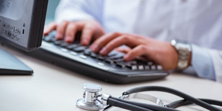 Una persona che indossa un camice bianco digita su una tastiera con uno stetoscopio posizionato sulla scrivania accanto a lei. La scena suggerisce un professionista medico che lavora su un computer, probabilmente gestendo le informazioni dei pazienti o conducendo ricerche all'interno del sistema sanitario portoghese.