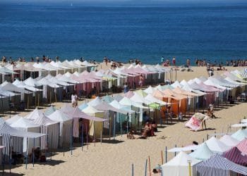 Un'affollata spiaggia sabbiosa è con bagnini che garantiscono gli interventi di soccorso sulle spiagge portoghesi. Le persone si rilassano sotto le tende e si godono il sole. Sullo sfondo si estende l'oceano azzurro e limpido, con poche barche e colline lontane visibili all'orizzonte.