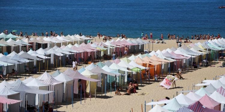 Un'affollata spiaggia sabbiosa è con bagnini che garantiscono gli interventi di soccorso sulle spiagge portoghesi. Le persone si rilassano sotto le tende e si godono il sole. Sullo sfondo si estende l'oceano azzurro e limpido, con poche barche e colline lontane visibili all'orizzonte.