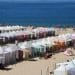 Un'affollata spiaggia sabbiosa è con bagnini che garantiscono gli interventi di soccorso sulle spiagge portoghesi. Le persone si rilassano sotto le tende e si godono il sole. Sullo sfondo si estende l'oceano azzurro e limpido, con poche barche e colline lontane visibili all'orizzonte.