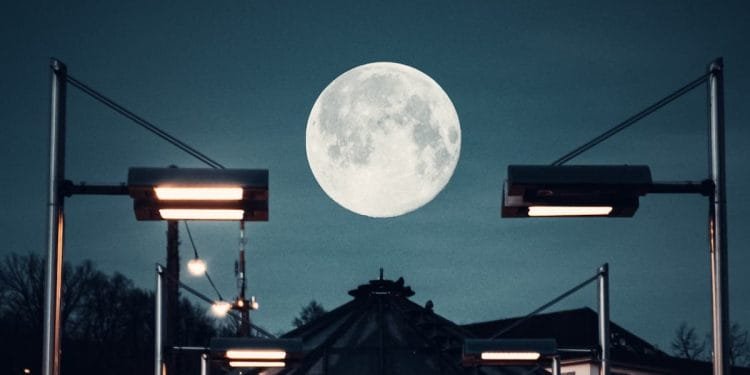Una grande superluna è ben visibile nel cielo notturno sopra una struttura urbana in Portogallo, incorniciata dai lampioni. Le luci illuminano il primo piano, proiettando una luce calda contro i toni freddi della sera e creando un'atmosfera serena e contrastante.