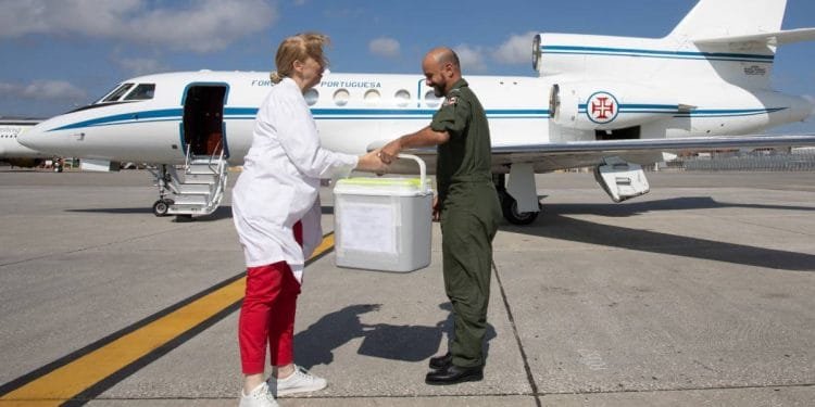 Una donna in camice bianco e pantaloni rossi porge un contenitore a un uomo in uniforme militare verde sulla pista dell'aeroporto, facilitando i trapianti tra il Portogallo e l'Italia. Sullo sfondo è parcheggiato un piccolo aeroplano bianco con la portiera aperta.
