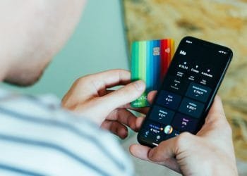 Una persona che tiene in una mano una carta di debito colorata e nell'altra uno smartphone su cui è visualizzata un'app di mobile banking. Lo schermo del telefono mostra i saldi dei conti, le transazioni, inclusi i bonifici bancari, e vari parametri finanziari. La persona, indossando una maglietta a righe, controlla il proprio numero cellulare per eventuali messaggi di conferma.