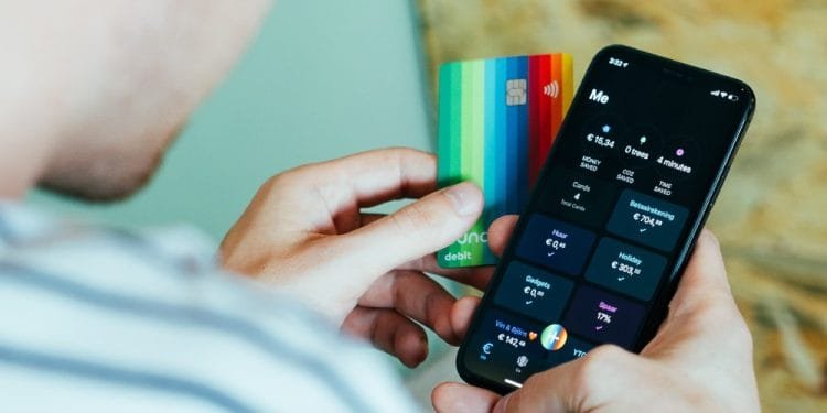 Una persona che tiene in una mano una carta di debito colorata e nell'altra uno smartphone su cui è visualizzata un'app di mobile banking. Lo schermo del telefono mostra i saldi dei conti, le transazioni, inclusi i bonifici bancari, e vari parametri finanziari. La persona, indossando una maglietta a righe, controlla il proprio numero cellulare per eventuali messaggi di conferma.