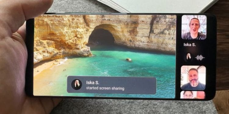 Una persona tiene in mano uno smartphone su cui è visualizzata una chat video su WhatsApp. La schermata principale mostra una scena di grotta e spiaggia, mentre una notifica dice "Iska S. ha avviato la condivisione schermo." Tre partecipanti sono in cornici più piccole sulla destra, con uno che parla attivamente.