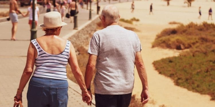 Una coppia di anziani, somigliante a pensionati italiani, cammina mano nella mano lungo una passerella vicino a una spiaggia sabbiosa. La donna indossa una canottiera a righe, pantaloncini blu e un cappello, mentre l'uomo indossa una maglietta grigia e pantaloncini scuri. Sullo sfondo si vedono persone che si godono la spiaggia e il lungomare.