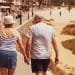 Una coppia di anziani, somigliante a pensionati italiani, cammina mano nella mano lungo una passerella vicino a una spiaggia sabbiosa. La donna indossa una canottiera a righe, pantaloncini blu e un cappello, mentre l'uomo indossa una maglietta grigia e pantaloncini scuri. Sullo sfondo si vedono persone che si godono la spiaggia e il lungomare.