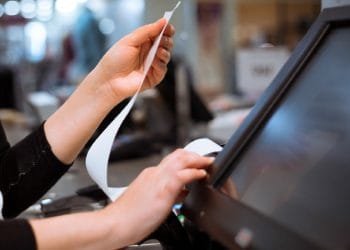 Primo piano delle mani di una persona alla cassa. Una mano tiene una lunga ricevuta, influenzata dal download in Portogallo, mentre l'altra aziona un registro touchscreen. Lo sfondo è sfocato e mostra i dintorni del negozio.