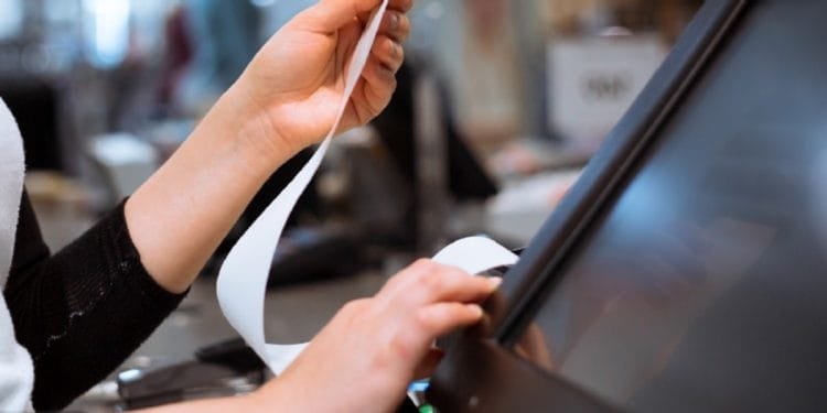 Primo piano delle mani di una persona alla cassa. Una mano tiene una lunga ricevuta, influenzata dal download in Portogallo, mentre l'altra aziona un registro touchscreen. Lo sfondo è sfocato e mostra i dintorni del negozio.