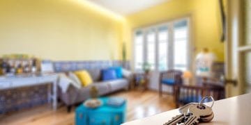 Un soggiorno accogliente e luminoso con pareti gialle e pavimento in legno. La camera dispone di un comodo divano, un tavolino e oggetti di arredamento che riflettono il mercato immobiliare portoghese. In primo piano, un mazzo di chiavi giace su una superficie bianca, con il focus sui tasti.