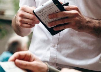 Primo piano di un cameriere in camicia bianca con in mano un blocco note e una penna, mentre prende un ordine da un cliente in un ambiente all'aperto. Il taccuino contiene appunti scritti a mano, con “portogallo prezzi” scarabocchiato in alto. La mano di un'altra persona che tiene un menu o un foglio è visibile in primo piano.