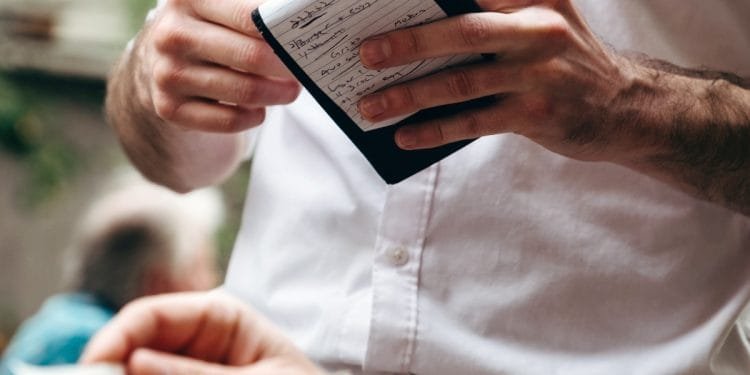 Primo piano di un cameriere in camicia bianca con in mano un blocco note e una penna, mentre prende un ordine da un cliente in un ambiente all'aperto. Il taccuino contiene appunti scritti a mano, con “portogallo prezzi” scarabocchiato in alto. La mano di un'altra persona che tiene un menu o un foglio è visibile in primo piano.