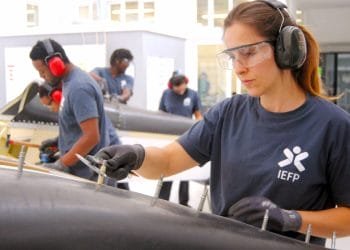 Una donna che indossa una maglietta "IEFP", a simboleggiare la sua partecipazione al programma IEFP ricerca lavoro Portogallo. Indossa occhiali protettivi, protezioni per le orecchie e guanti lavora meticolosamente su un oggetto in un'officina. Sullo sfondo, altre tre persone, anch'esse con indumenti protettivi e magliette simili, si concentrano sui rispettivi compiti.