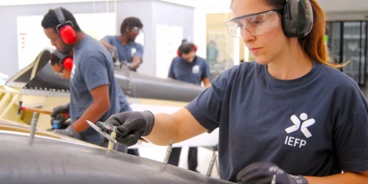 Una donna che indossa una maglietta "IEFP", a simboleggiare la sua partecipazione al programma IEFP ricerca lavoro Portogallo. Indossa occhiali protettivi, protezioni per le orecchie e guanti lavora meticolosamente su un oggetto in un'officina. Sullo sfondo, altre tre persone, anch'esse con indumenti protettivi e magliette simili, si concentrano sui rispettivi compiti.