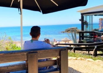 Una persona siede su una panchina sotto un grande ombrellone, guardando una spiaggia con un mare cristallino. Alcune persone sono sulla spiaggia e in acqua. A destra si trova un edificio moderno con grandi finestre dove si potrebbe aprire attività portogallo. Il sole splende, suggerendo una giornata calda.