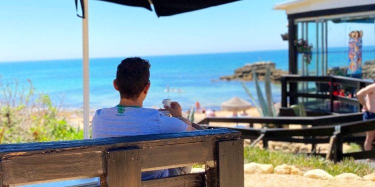 Una persona siede su una panchina sotto un grande ombrellone, guardando una spiaggia con un mare cristallino. Alcune persone sono sulla spiaggia e in acqua. A destra si trova un edificio moderno con grandi finestre dove si potrebbe aprire attività portogallo. Il sole splende, suggerendo una giornata calda.