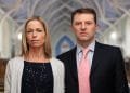 Una donna e un uomo stanno fianco a fianco in una chiesa. La donna, con i capelli biondi lunghi fino alle spalle, indossa un cardigan chiaro sopra un top scuro. L'uomo indossa un abito scuro con una cravatta rosa chiaro. Entrambi hanno espressioni serie e guardano direttamente la telecamera, ricordando figure legate al caso Maddie McCann.