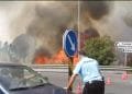 Un agente di polizia si sporge verso un'auto su una strada con fumo pesante e fiamme visibili sullo sfondo. Sono allestiti coni stradali e i segnali nelle vicinanze indicano le direzioni per Funchal, Ribeira Brava e Calheta. Il cielo è pieno di fumo denso causato dal violento incendio in Portogallo.