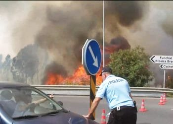 Un agente di polizia si sporge verso un'auto su una strada con fumo pesante e fiamme visibili sullo sfondo. Sono allestiti coni stradali e i segnali nelle vicinanze indicano le direzioni per Funchal, Ribeira Brava e Calheta. Il cielo è pieno di fumo denso causato dal violento incendio in Portogallo.