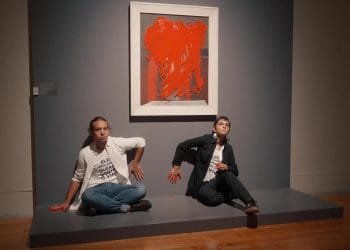 Due individui sono seduti su una piattaforma in una galleria. Entrambi hanno la mano destra, imbrattata di vernice rossa, premuta contro il muro. Dietro di loro c'è un dipinto astratto incorniciato con audaci tratti rossi. La galleria ha pavimenti in legno e un muro grigio tenue, che evoca lo spirito degli attivisti di Lisbona.