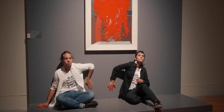 Due individui sono seduti su una piattaforma in una galleria. Entrambi hanno la mano destra, imbrattata di vernice rossa, premuta contro il muro. Dietro di loro c'è un dipinto astratto incorniciato con audaci tratti rossi. La galleria ha pavimenti in legno e un muro grigio tenue, che evoca lo spirito degli attivisti di Lisbona.