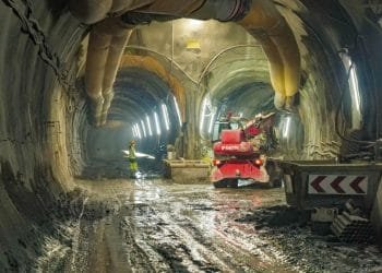 Un operaio edile in abiti ad alta visibilità si trova in un tunnel fangoso e poco illuminato, illuminato da luci fluorescenti. Macchinari pesanti, tra cui un veicolo rosso contrassegnato con "MANITOU", operano sullo sfondo tra tubi e materiali da costruzione per le nuove linee metropolitane di Porto.
