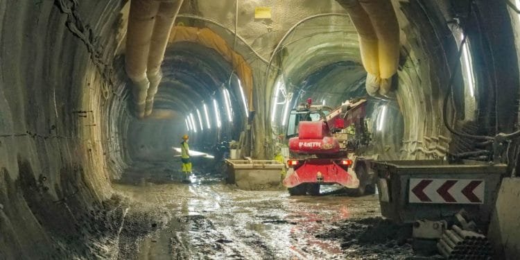 Un operaio edile in abiti ad alta visibilità si trova in un tunnel fangoso e poco illuminato, illuminato da luci fluorescenti. Macchinari pesanti, tra cui un veicolo rosso contrassegnato con "MANITOU", operano sullo sfondo tra tubi e materiali da costruzione per le nuove linee metropolitane di Porto.