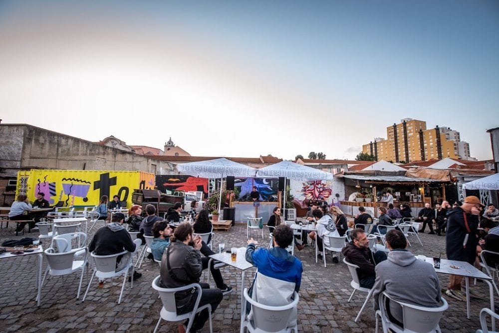 Un caffè all'aperto negli spazi culturali Lisbona con persone sedute a tavoli e sedie bianchi su un patio acciottolato. Murales colorati decorano le pareti di sfondo e gli edifici sono visibili in lontananza sotto un cielo limpido. Alcuni avventori parlano, mentre altri si godono un drink.