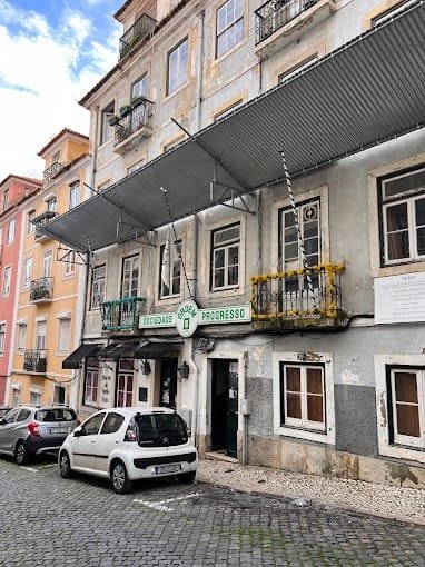 Una vista sulla strada di un edificio a più piani esposto alle intemperie con un cartello con la scritta "Sociedade Progresso" sopra l'ingresso. L'edificio, parte di uno dei vivaci spazi culturali di Lisbona, è dotato di balconi, alcuni con cesti di fiori. Lungo la strada acciottolata sono parcheggiate diverse auto. Gli edifici vicini sono colorati e simili nello stile.