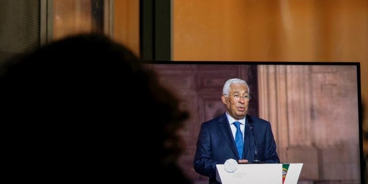 Una persona sta guardando uno schermo televisivo su cui si vede un uomo in giacca e cravatta che tiene un discorso. L'oratore, che ha i capelli bianchi, è in piedi su un podio con uno stemma e una piccola bandiera nell'angolo. Lo sfondo, che con le sue sfumature arancioni e marroni conferisce un'aria di serenità, sussurra l'appello del Portogallo ai pensionati.