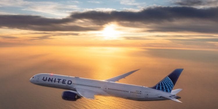 Un aereo passeggeri della United Airlines sta volando sopra uno specchio d'acqua durante il tramonto, probabilmente su un volo da o per l'aeroporto di Faro. Il cielo è pieno di morbide nuvole mentre il sole proietta un bagliore dorato sulla scena, illuminando l'intera lunghezza e il marchio dell'aereo dalla sua vista laterale.