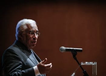 Un uomo in abito e occhiali parla al microfono su un'asta, gesticolando con la mano destra come se si rivolgesse a un pubblico in una stanza scarsamente illuminata con uno sfondo marrone. Sembra che Antonio Costa abbia appena annunciato le sue dimissioni.