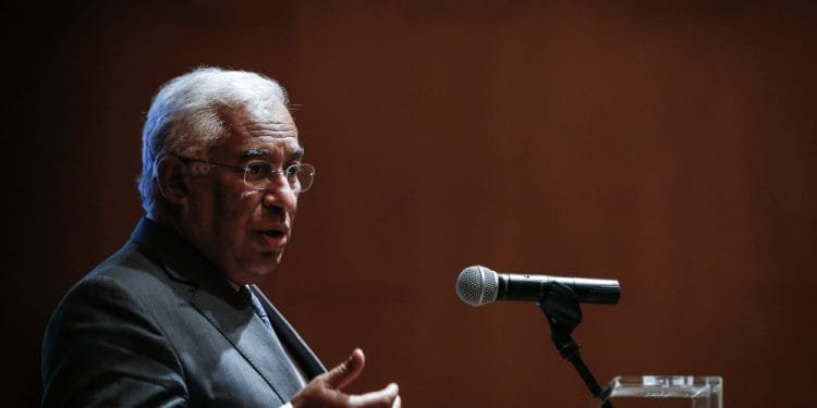 Un uomo in abito e occhiali parla al microfono su un'asta, gesticolando con la mano destra come se si rivolgesse a un pubblico in una stanza scarsamente illuminata con uno sfondo marrone. Sembra che Antonio Costa abbia appena annunciato le sue dimissioni.