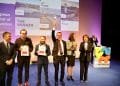 Un gruppo di persone si trova su un palco durante una cerimonia di premiazione. Un uomo al centro alza un trofeo, celebrando Lisbona Capitale Europea Innovazione. Altri hanno dei certificati e sullo sfondo c'è uno schermo che mostra Lisbona, Varsavia e Lviv.