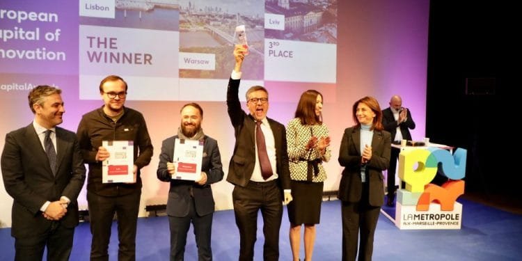Un gruppo di persone si trova su un palco durante una cerimonia di premiazione. Un uomo al centro alza un trofeo, celebrando Lisbona Capitale Europea Innovazione. Altri hanno dei certificati e sullo sfondo c'è uno schermo che mostra Lisbona, Varsavia e Lviv.