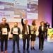 Un gruppo di persone si trova su un palco durante una cerimonia di premiazione. Un uomo al centro alza un trofeo, celebrando Lisbona Capitale Europea Innovazione. Altri hanno dei certificati e sullo sfondo c'è uno schermo che mostra Lisbona, Varsavia e Lviv.